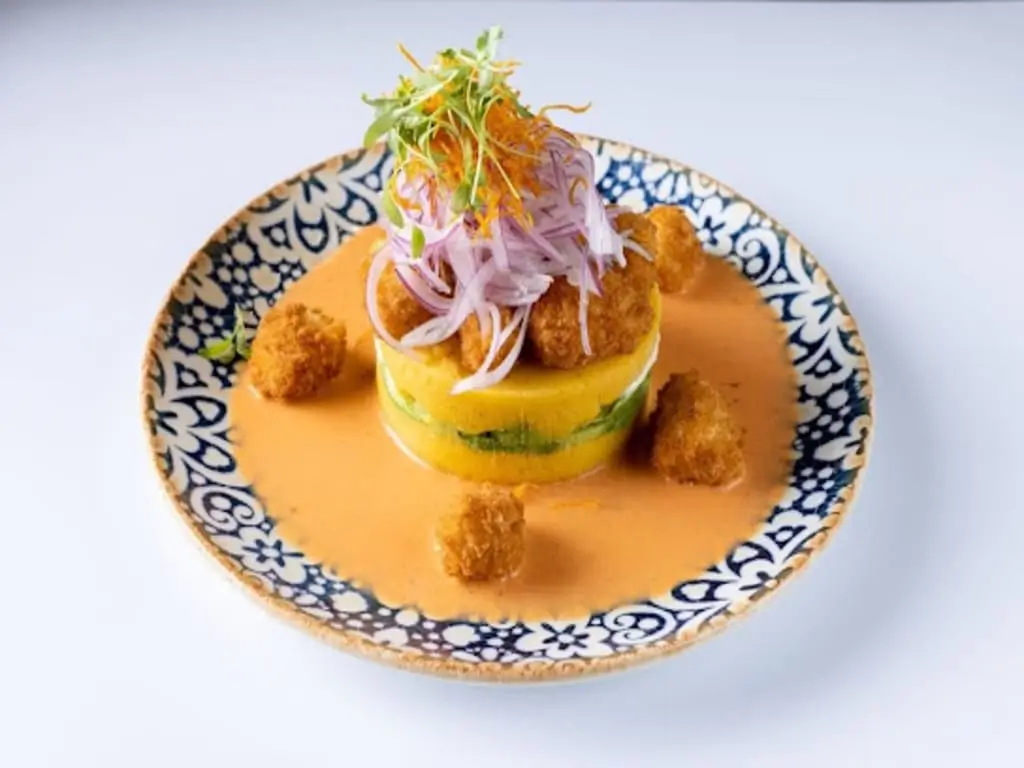 Peruvian causa acevichada with fried seafood and creamy sauce on decorative plate.