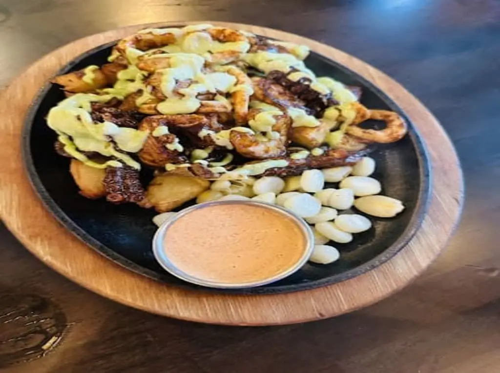 Grilled Peruvian seafood platter with octopus, calamari, huancaína sauce, Peruvian corn, and dipping sauce on a wooden tray. - Manta Restaurant